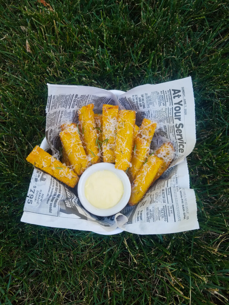 Parmesan grated over baked polenta fries in a newspaper lined basket with a garlic aioli dip