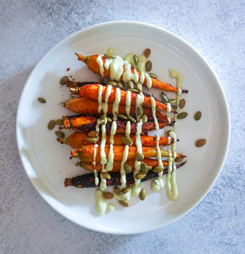 Plate of harissa roasted carrots with an avocado yogurt sauce drizzled on top and pepitos.